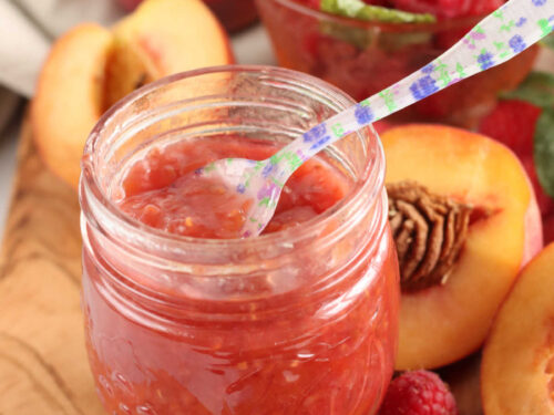 Jar of raspberry peach jam on wooden cutting board, floral spoon in jar.