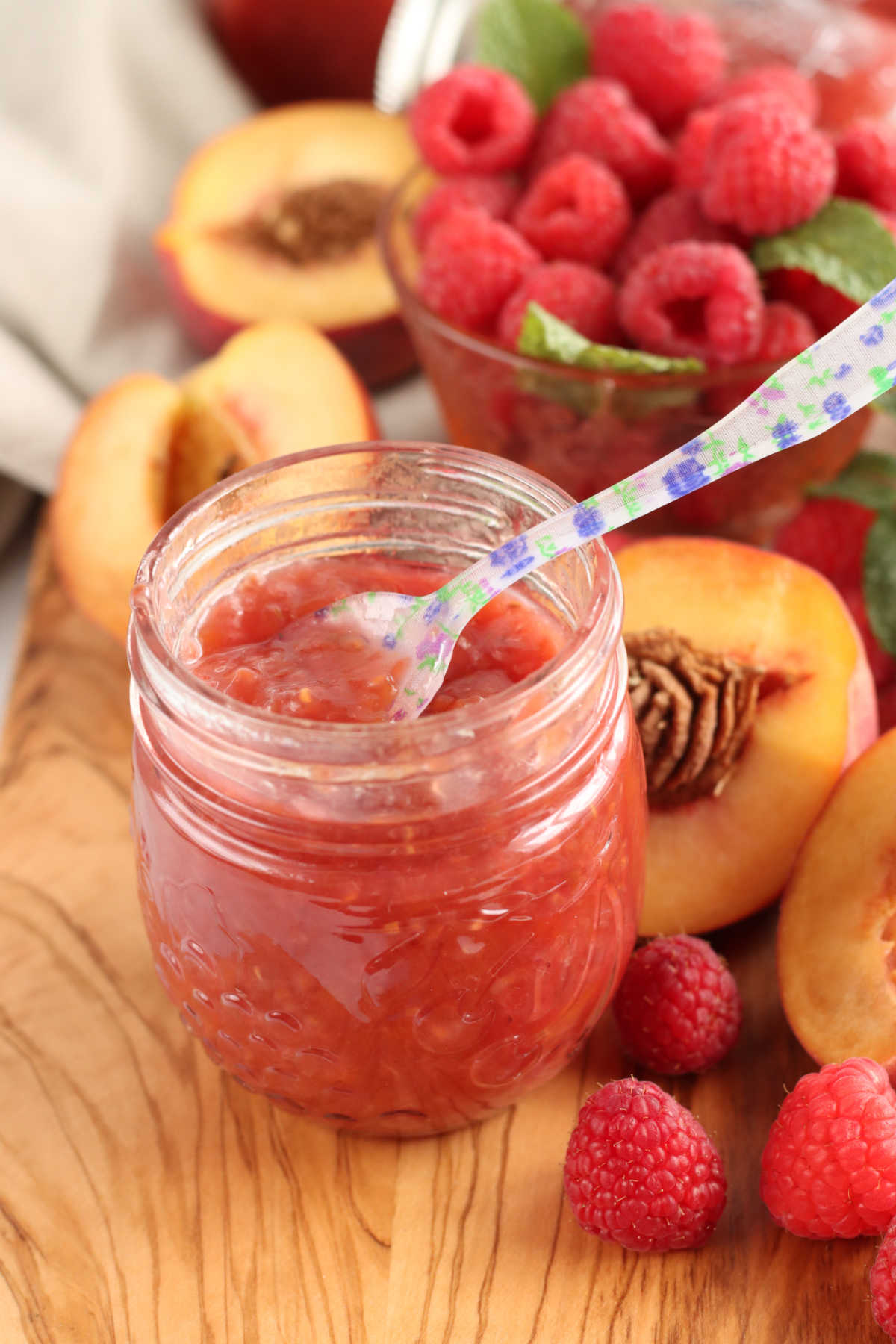 Clear jar of peach raspberry jam on wooden cutting board, floral spoon in jar, berries around.