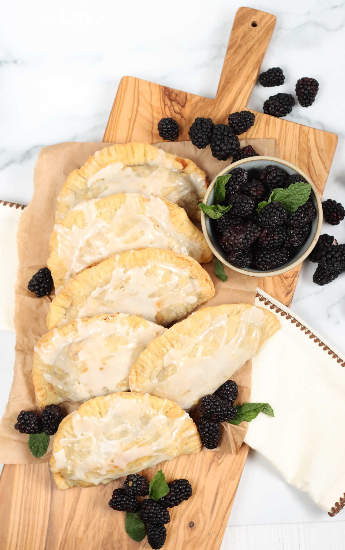 Blackberry hand pies with glaze on a wooden cutting board, loose berries around them.