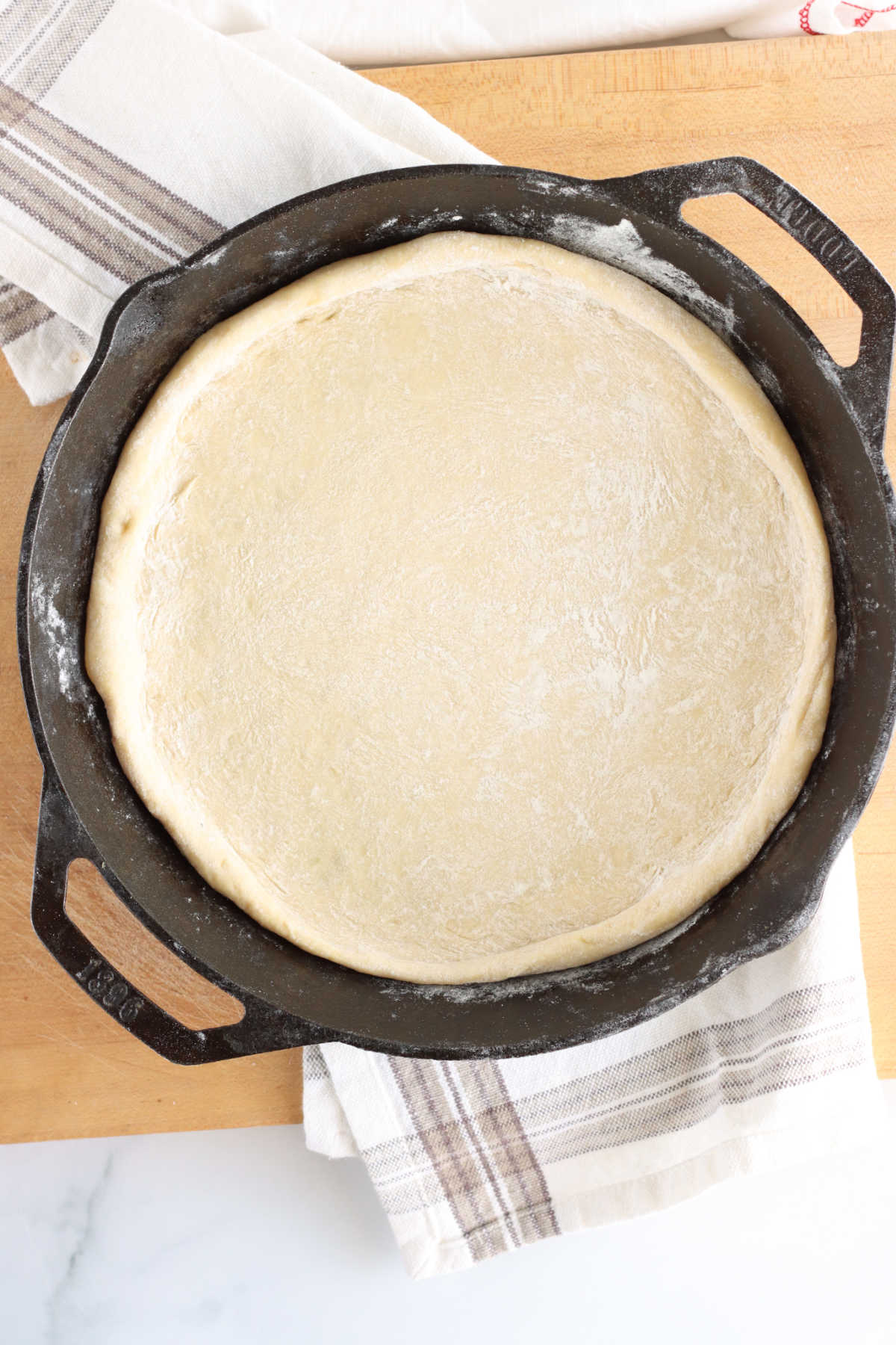 Pizza dough rising in cast iron skillet on wooden cutting board, kitchen towel underneath.