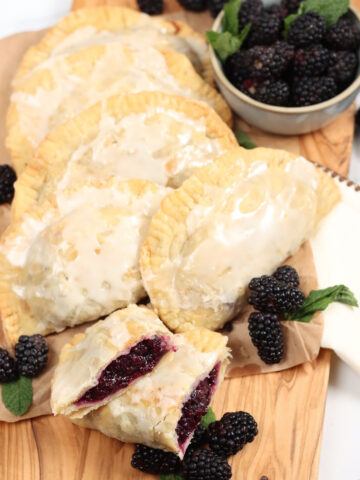 Blackberry hand pies on wooden cutting board, loose blackberries around.