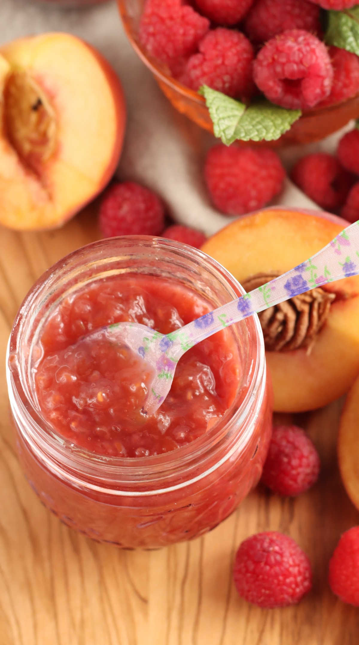 Close up of glass jar with peach raspberry jam on wooden cutting board, fresh fruit in background.