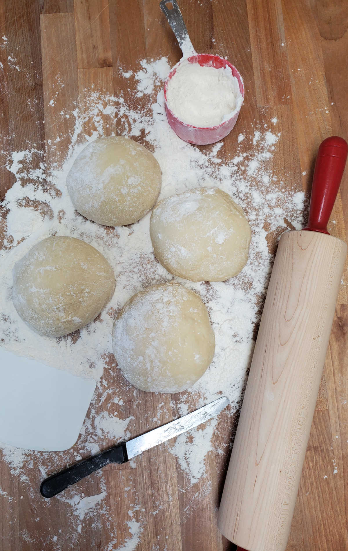 Four rounds of pizza dough rising on butcher block, red handled rolling pin to side.