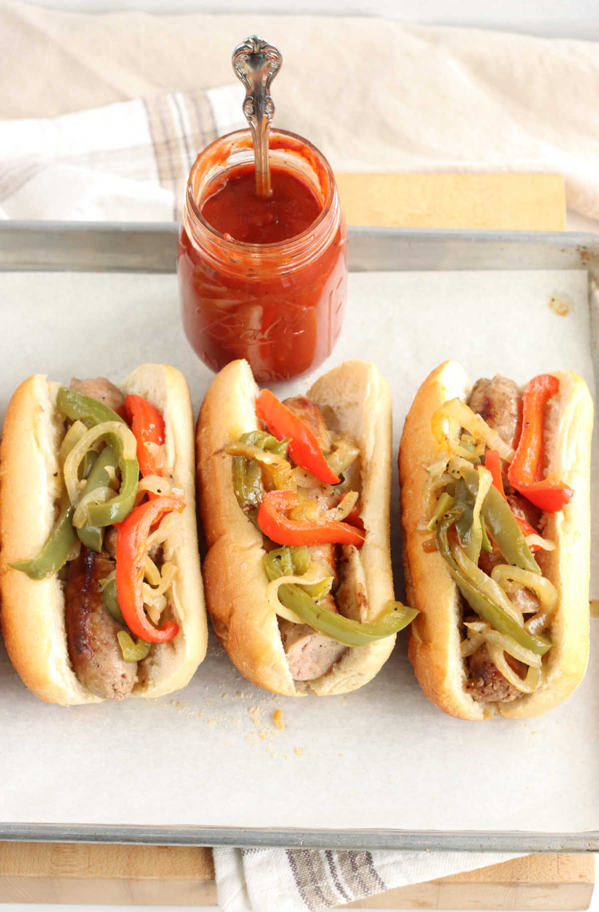 Sausage and peppers on Hoagie rolls on sheet pan with parchment paper, jar of sauce with spoon.