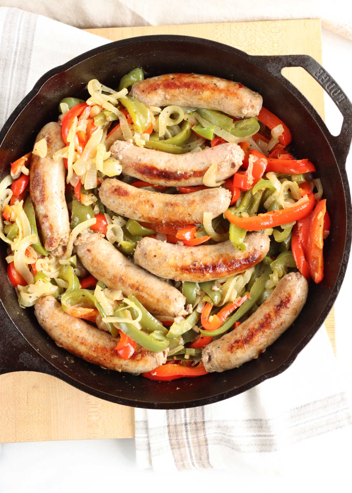 Browned sausage links, peppers, onions in cast iron skillet on wooden cutting board.