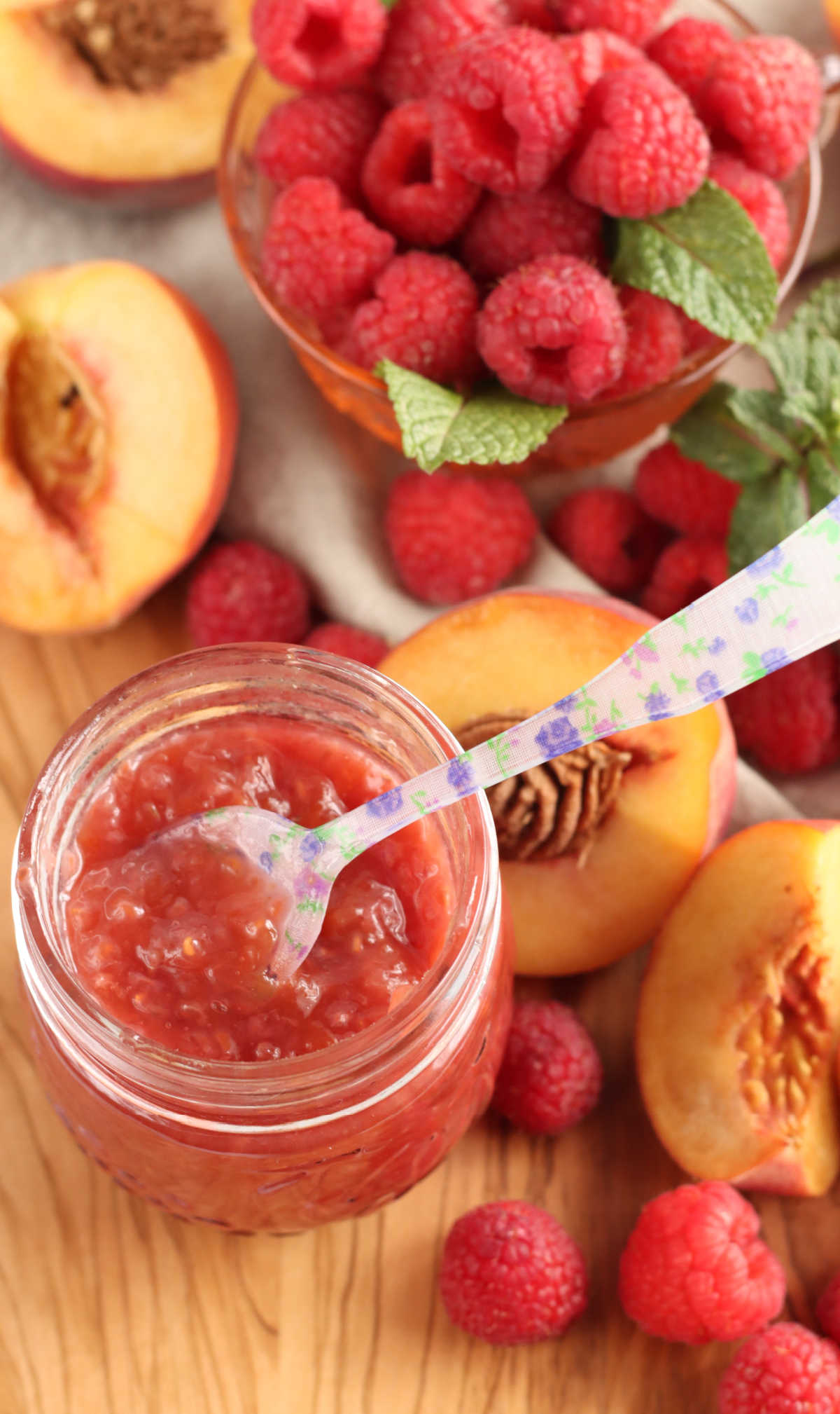 Glass jar of peach raspberry jam, floral spoon in jar, peaches and raspberries on cutting board.