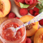Glass jar of raspberry peach jam on wooden cutting board, fresh raspberries and peaches around.