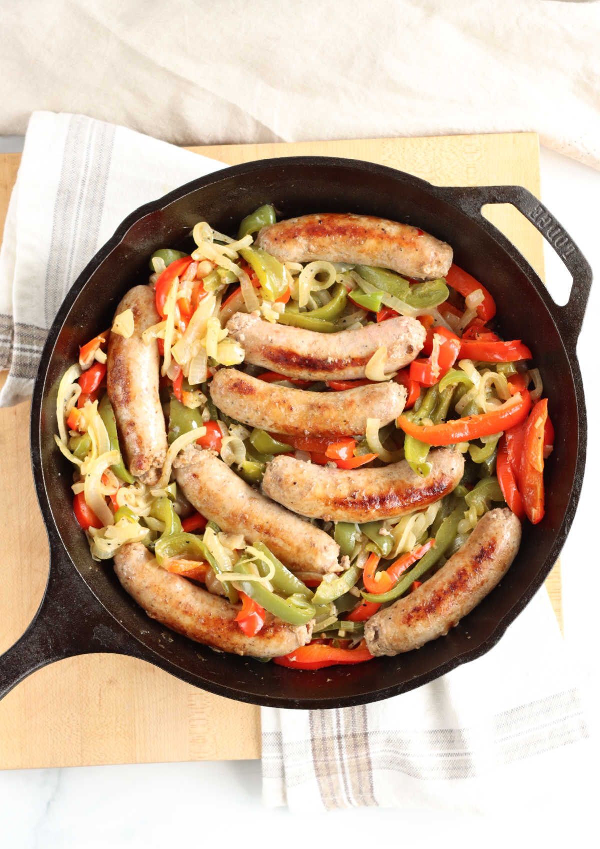 Sausage links, onions, green and red Bell peppers in cast iron skillet on wooden cutting board.