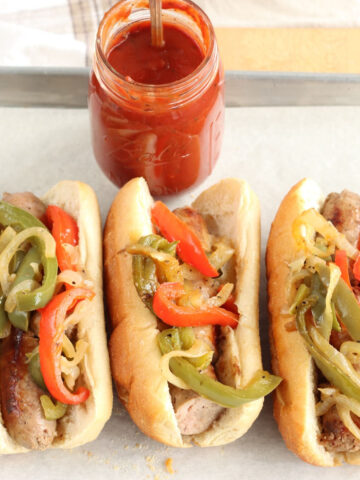 Sausage and pepper subs on Hoagie rolls on half sheet pan, jar of marinara sauce with spoon in background.
