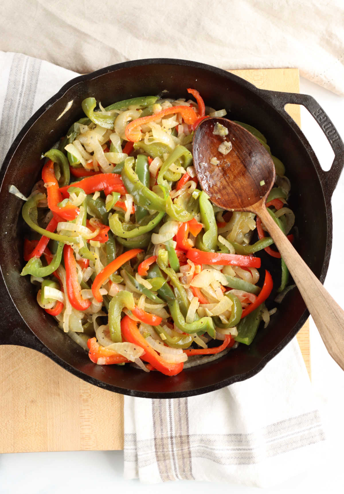 Red and green Bell peppers, onions in large cast iron skillet, wooden spoon in right of pan.