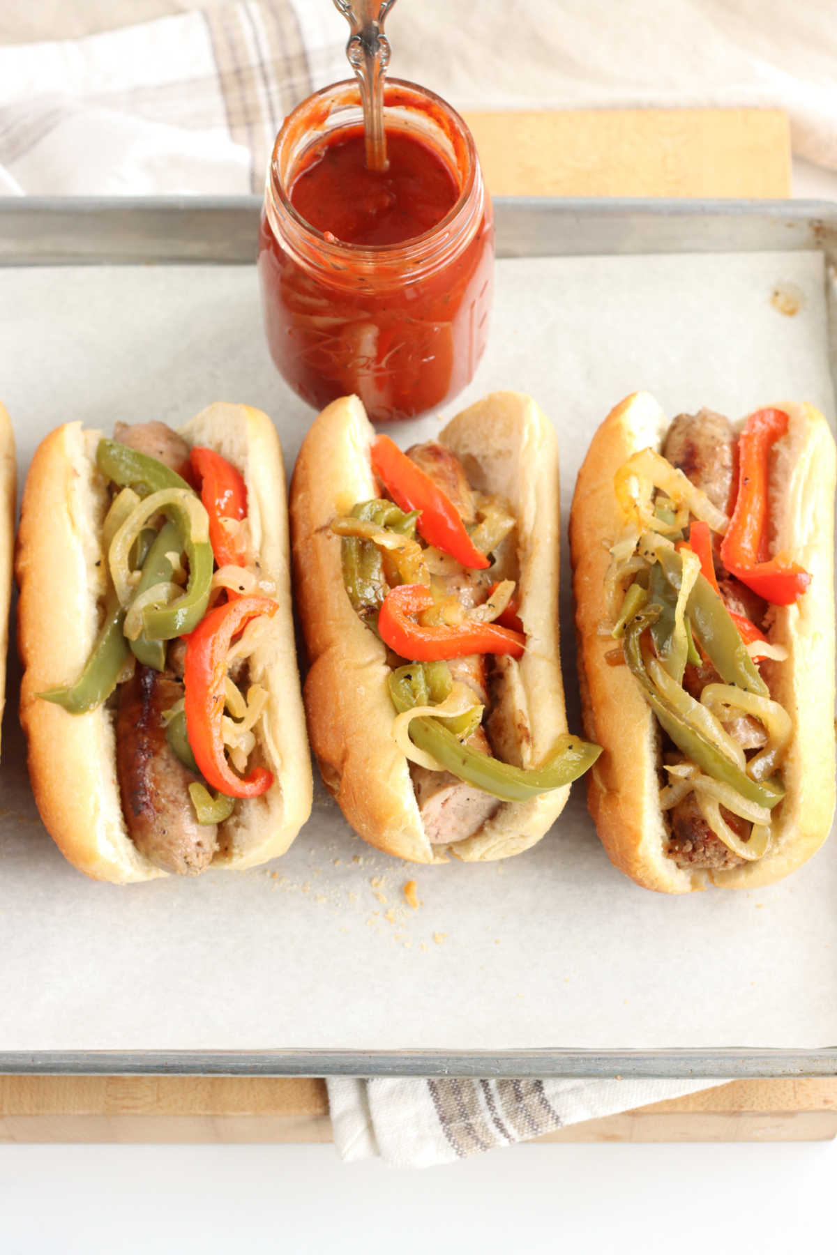 Hoagie rolls with sausage, peppers and onions on half sheet pan, jar of sauce in background.