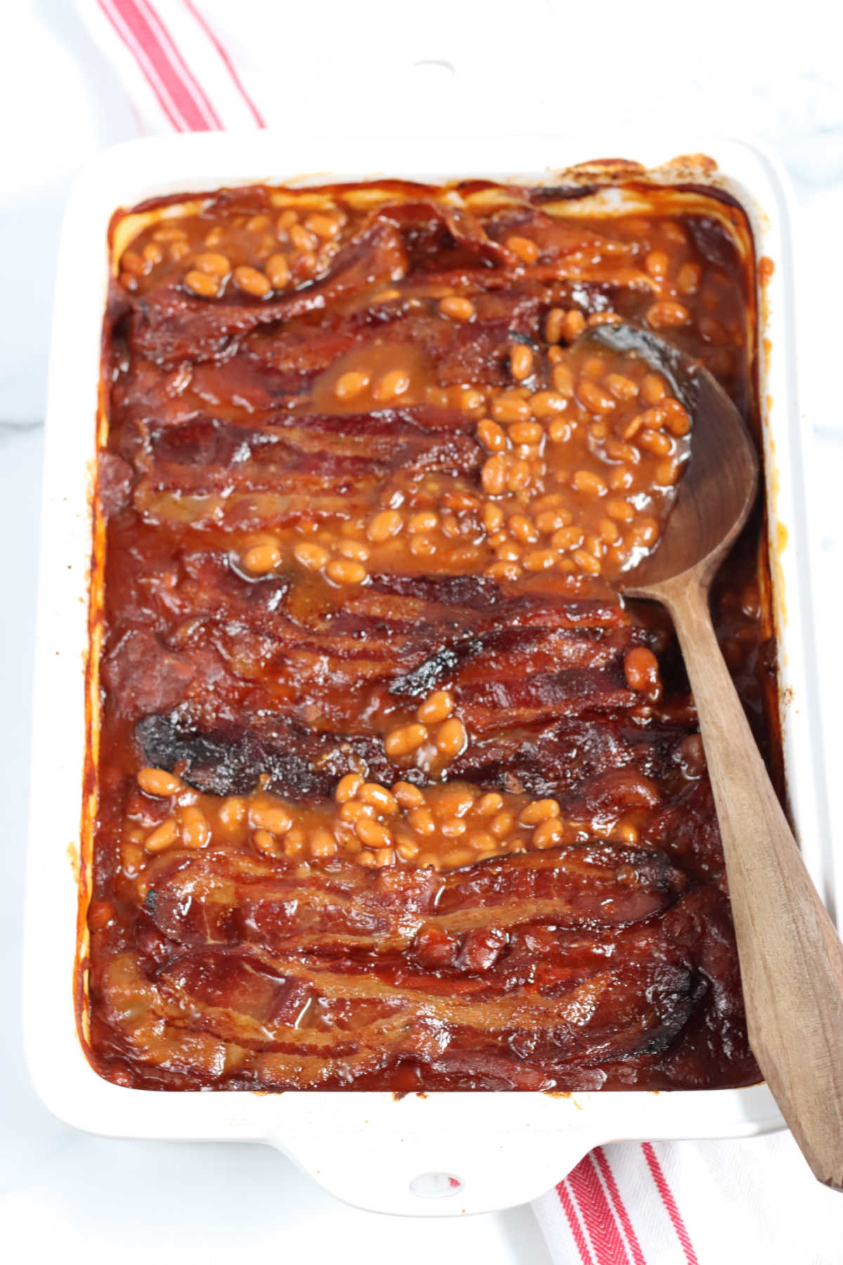 Baked beans topped with crispy bacon in white rectangle baking dish, wooden spoon in right of dish.