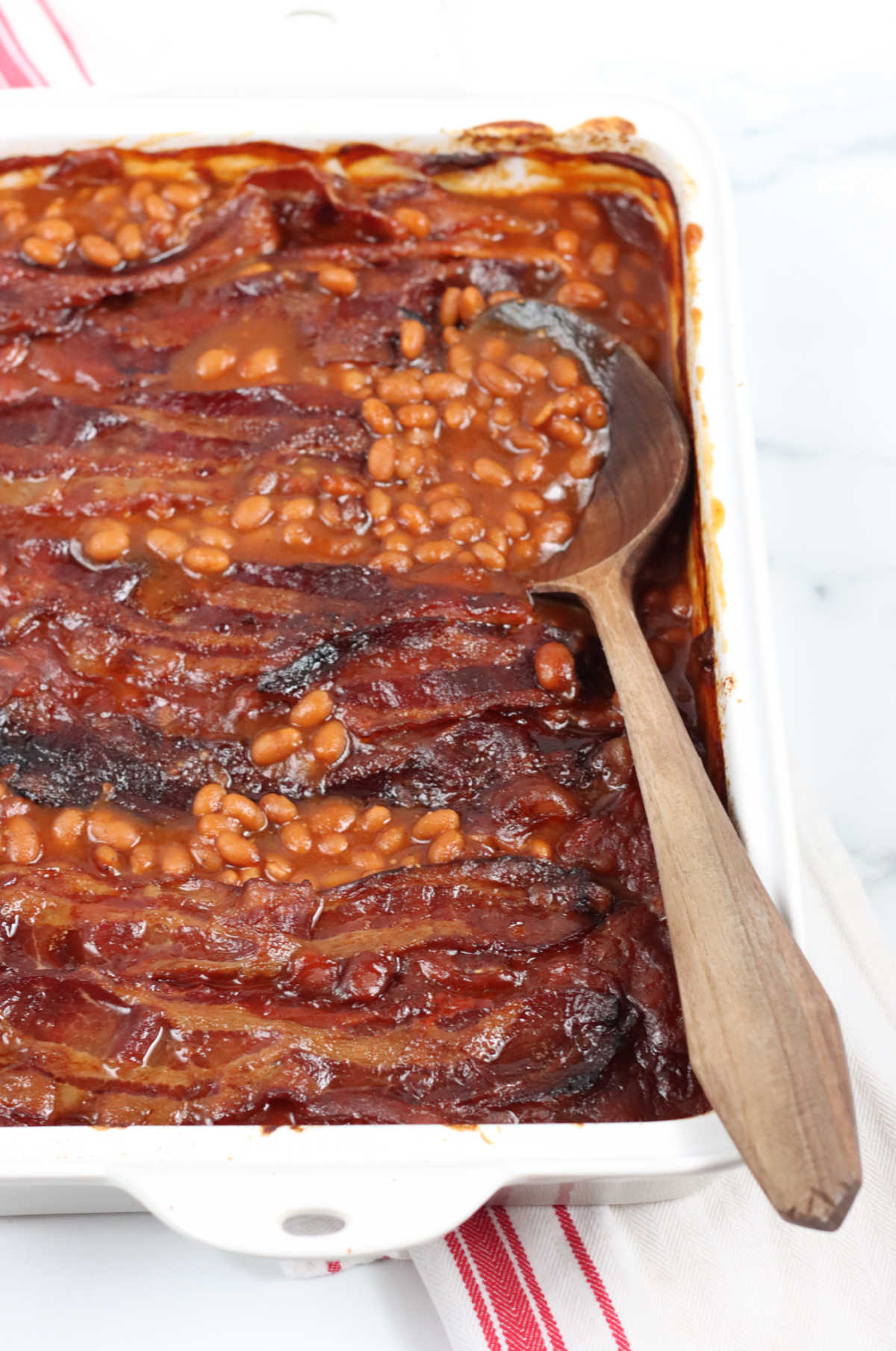 Baked beans in white rectangle baking dish, wooden spoon on right side of pan.