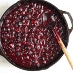 Cherry pie filling in large cast iron skillet on white marble, wooden spoon in pan.