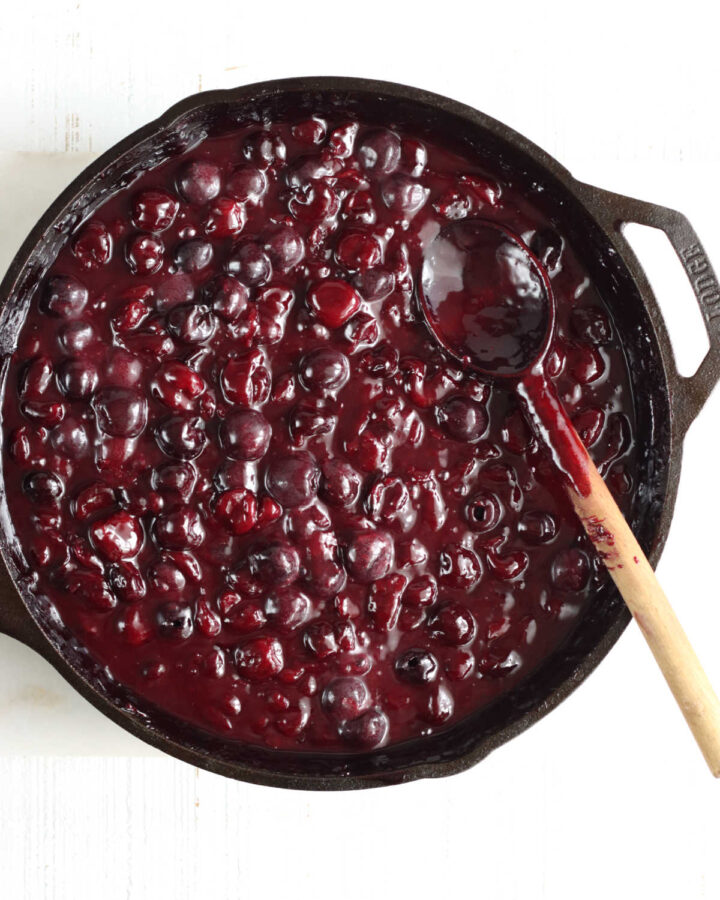 Cherry pie filling in large cast iron skillet on white marble, wooden spoon in pan.