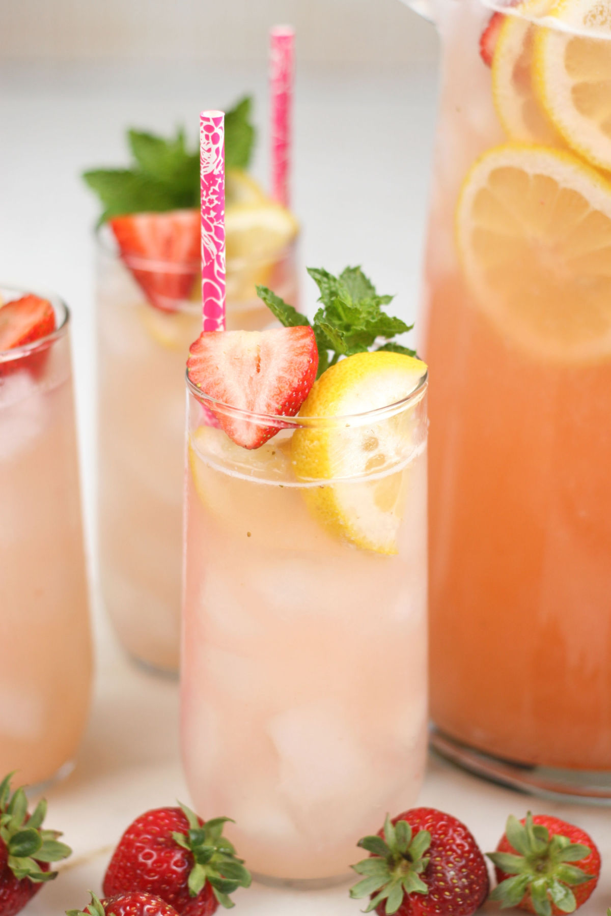 Clear cylinder glass of strawberry lemonade, fresh strawberry slices, ice cubes, lemon slices, fresh mint in glass.