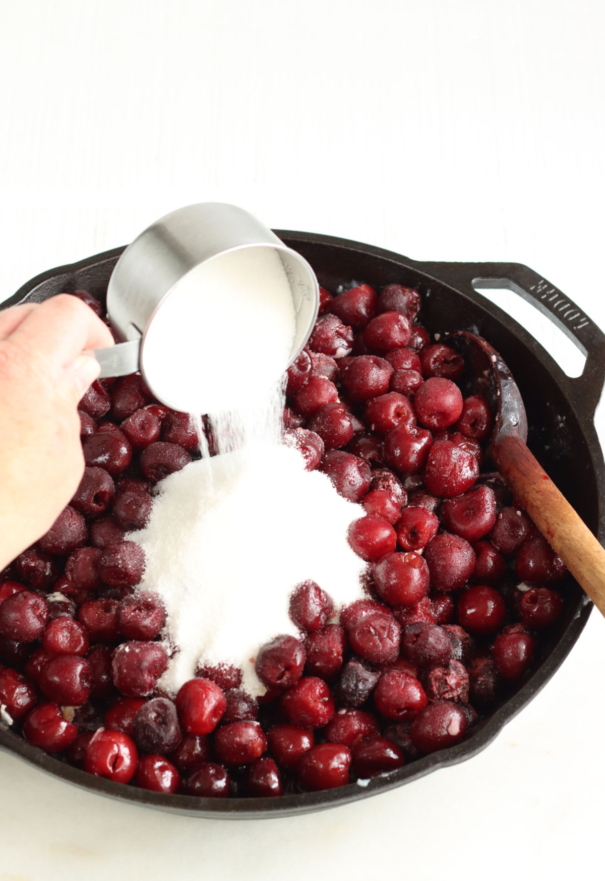 Pouring sugar from metal measuring cup into cherry pie filling in large cast iron skillet.