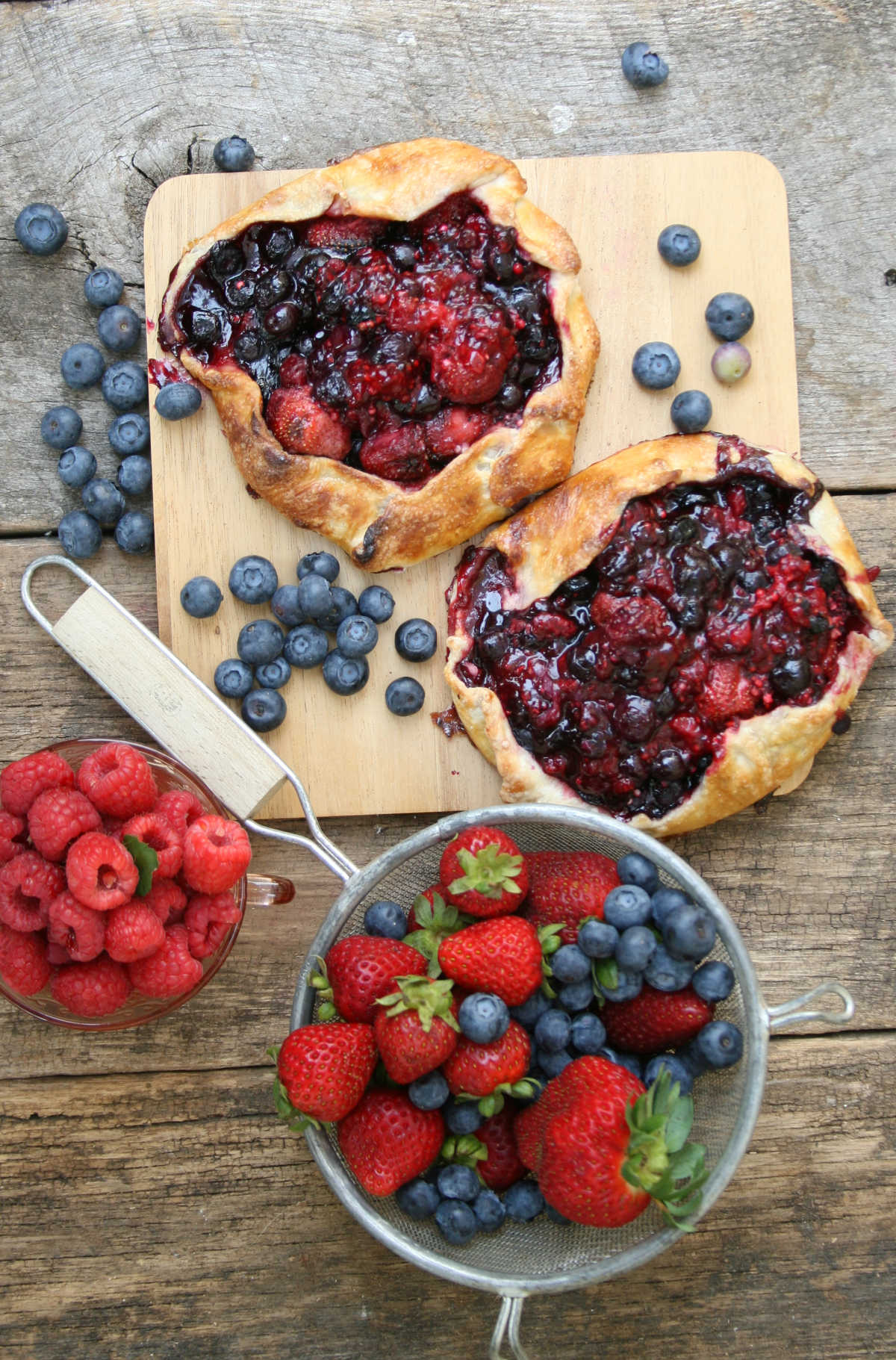Berry galettes on wooden cutting board, strawberries, blueberries, raspberries in metal strainer.