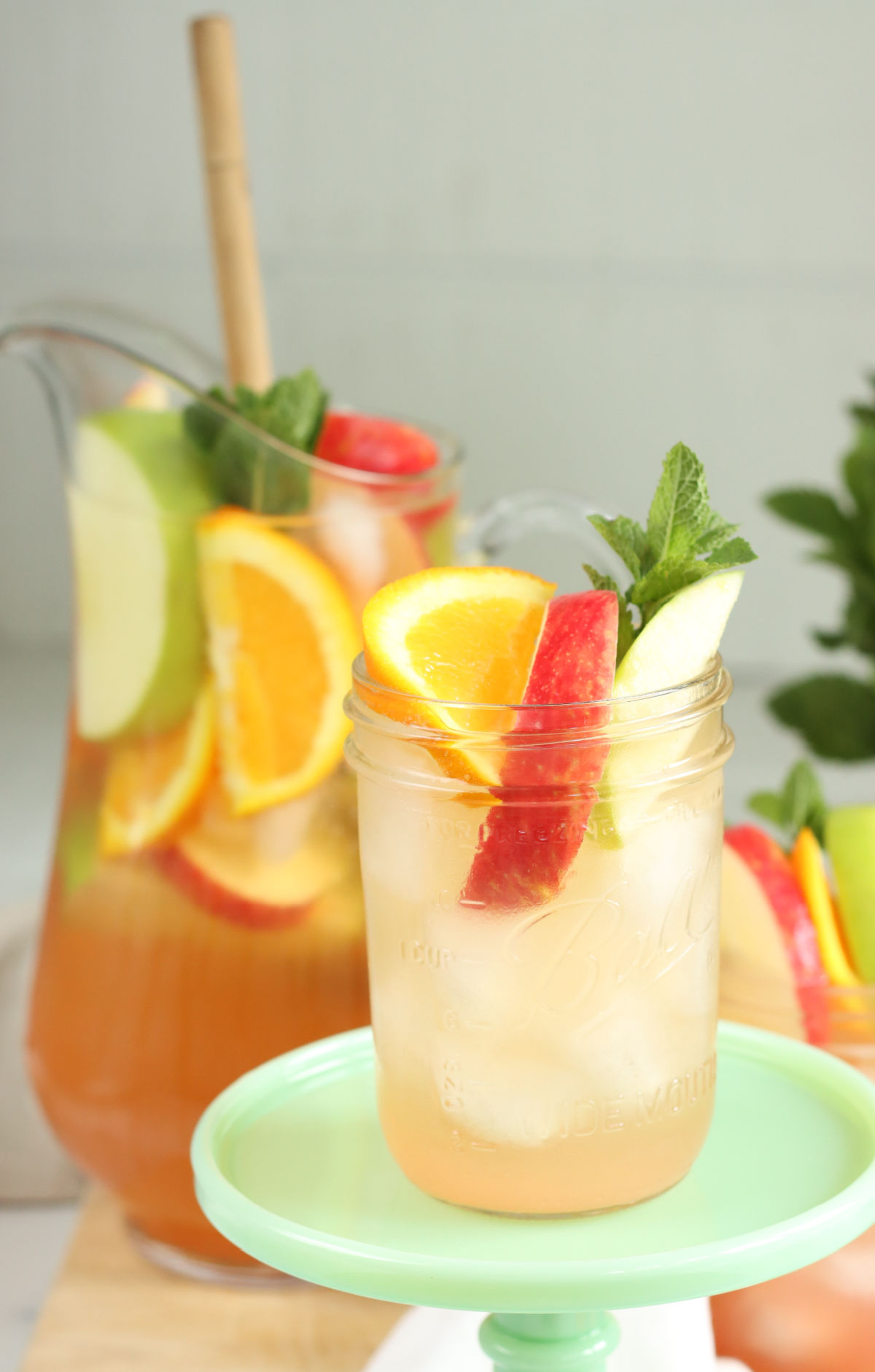 Close up of Pint Mason jar with sangria, apple and orange slices, pouring pitcher in background.