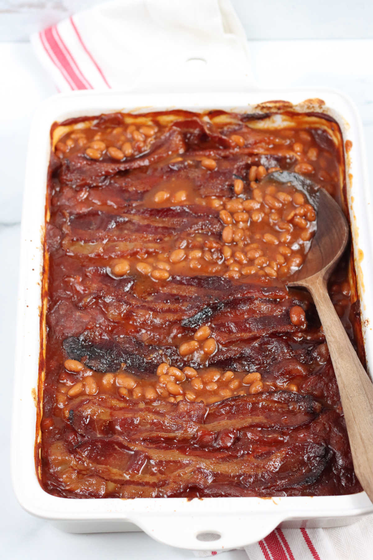 Baked beans in white rectangle baking dish, wooden spoon on right.
