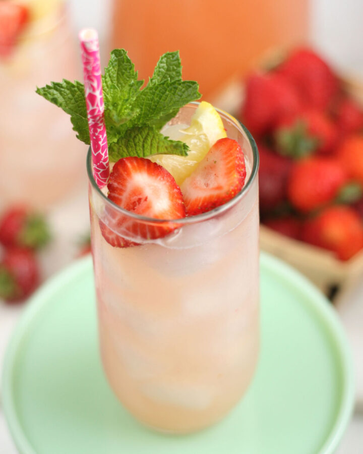 Glass of strawberry lemonade, fresh strawberries, lemon slices, fresh mint, raspberry paper straw in glass.