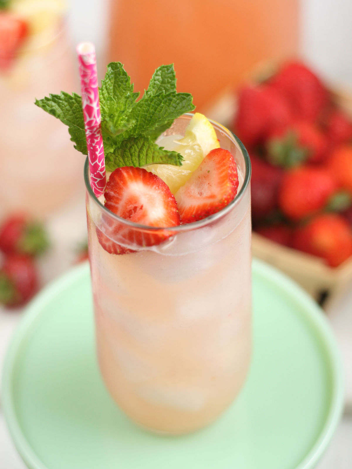 Glass of strawberry lemonade, fresh strawberries, lemon slices, fresh mint, raspberry paper straw in glass.