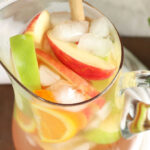 Glass pouring pitcher with white sangria, apple and orange slices, ice cubes.