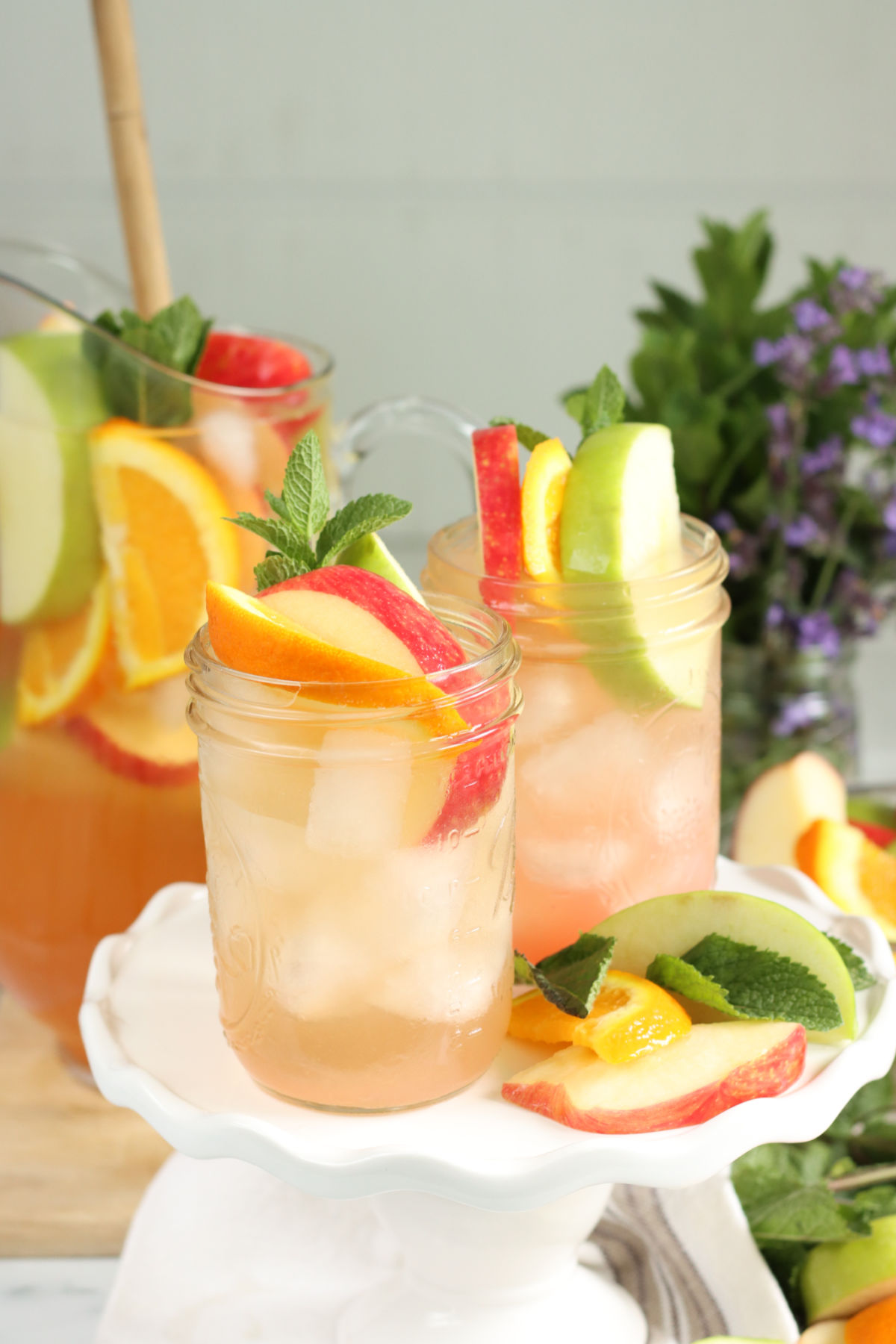 Two pint wide-mouth Mason jars with sangria, orange and apples slices, pouring pitcher in background.