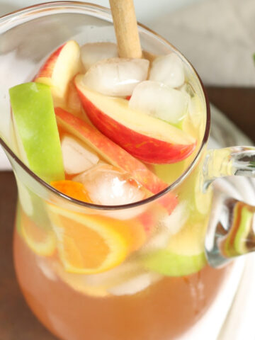 Clear glass pouring pitcher of white sangria with ice cubes, red and green apple slices, orange slices.
