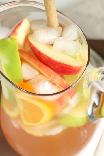 Clear glass pouring pitcher of white sangria with ice cubes, red and green apple slices, orange slices.