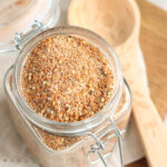 Square glass jar with BBQ spice rub, wooden tablespoon in background.