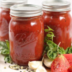 Mason jars of marinara sauce on wooden cutting board, tomato quarters, fresh herbs, whole garlic cloves.