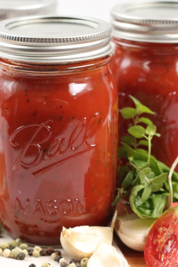 Mason jars of marinara sauce on wooden cutting board, tomato quarters, fresh herbs, whole garlic cloves.