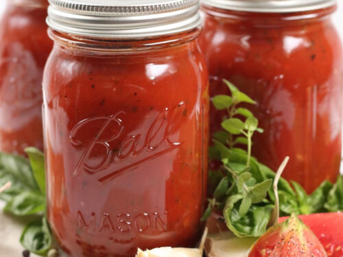 Mason jars of marinara sauce on wooden cutting board, tomato quarters, fresh herbs, whole garlic cloves.