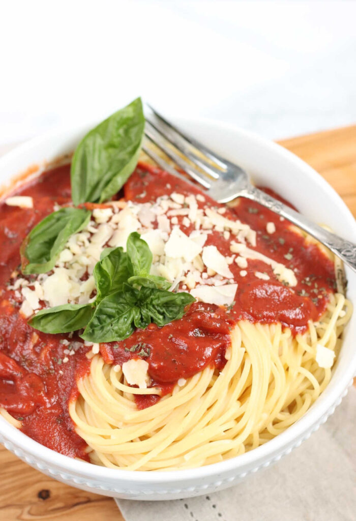 White bowl with spaghetti, marinara sauce, shaved Parmesan cheese, fresh basil.