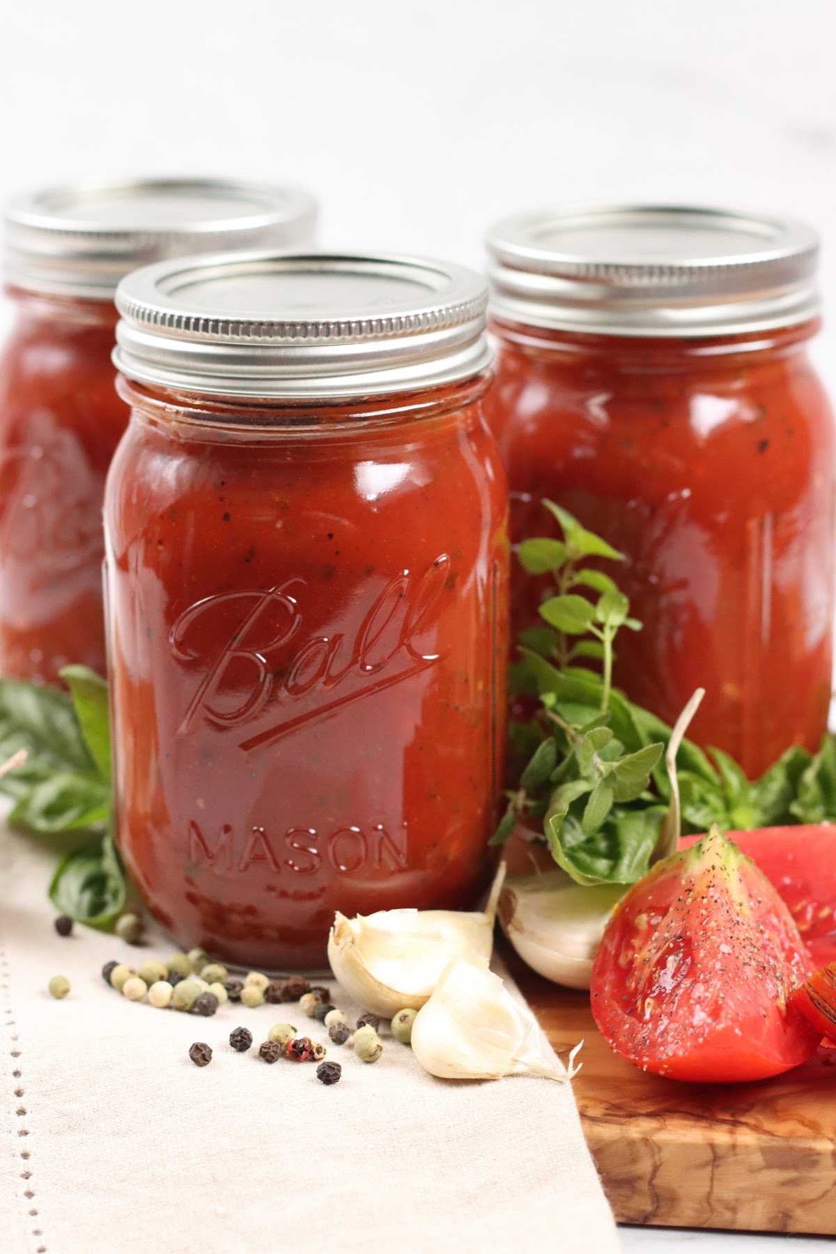 Pint Mason jars of marinara sauce on wooden cutting board, tomato quarters, whole garlic cloves and herbs.