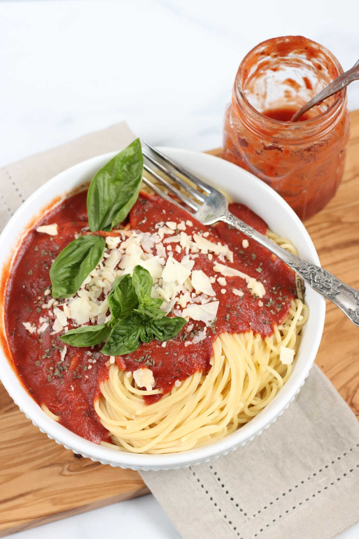 White bowl of spaghetti, marinara sauce, basil leaves, shaved Parmesan cheese, jar of sauce.