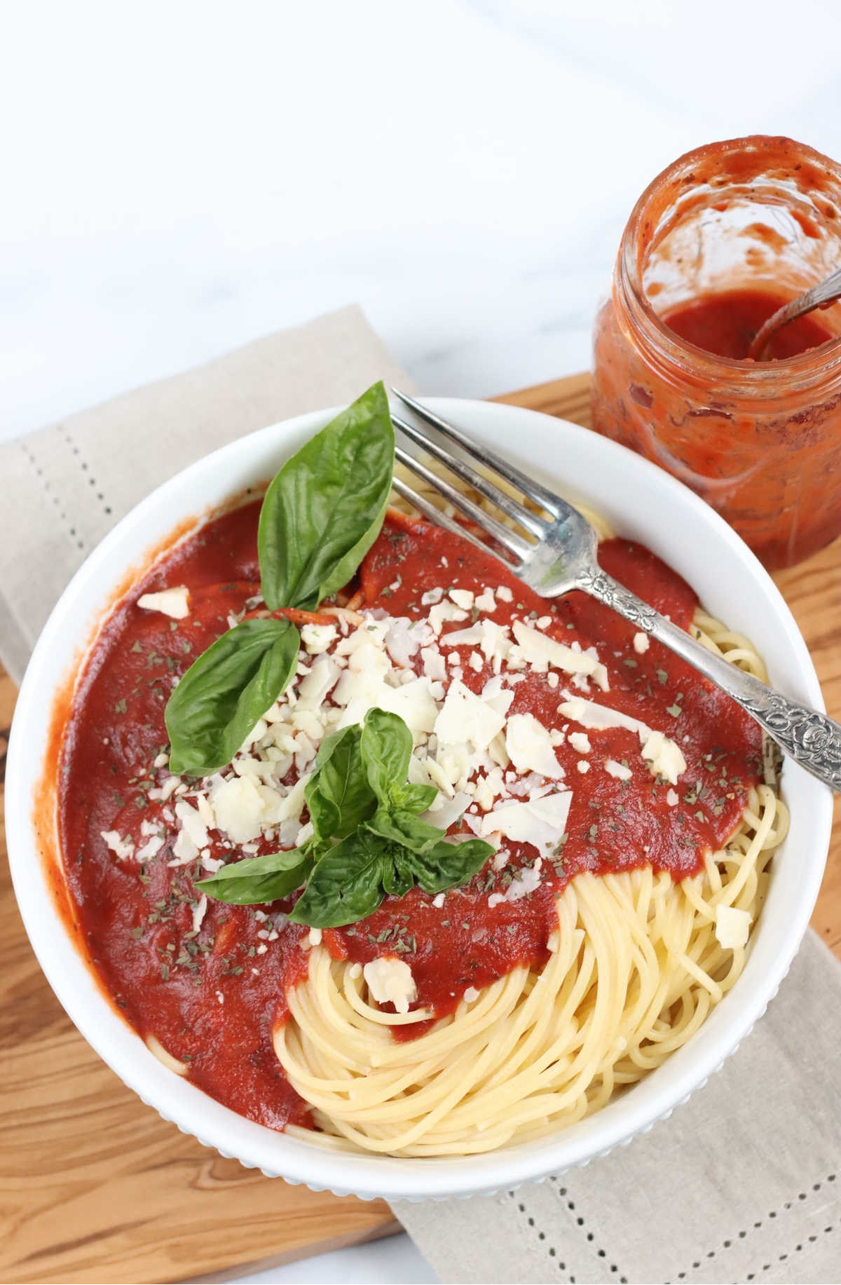 White bowl of spaghetti with marinara sauce, shaved Parmesan cheese, fresh basil, jar in background.