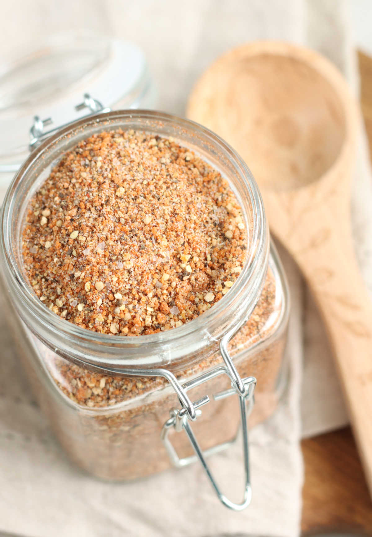 Square glass jar with BBQ spice rub, wooden tablespoon in background.