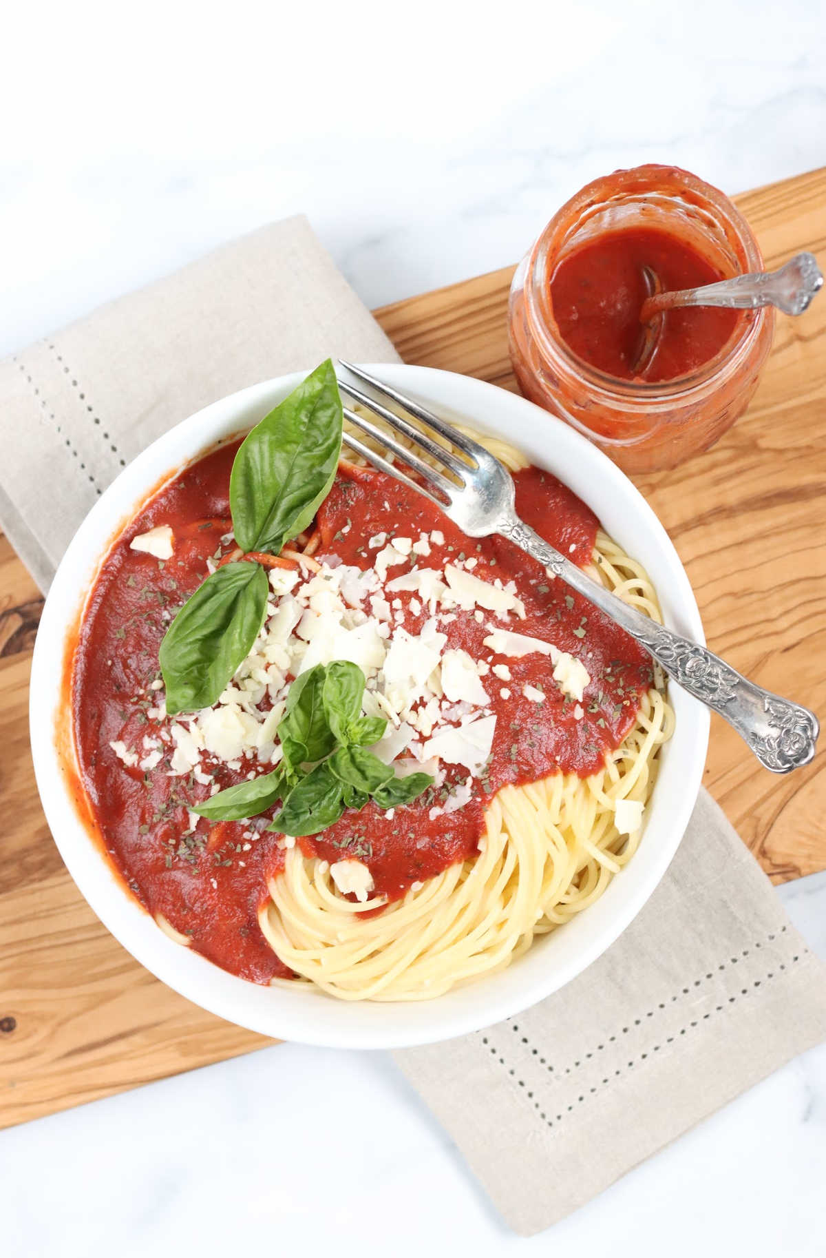 White bowl of spaghetti, marinara sauce, Parmesan cheese, fresh basil, jar of sauce on wooden cutting board.
