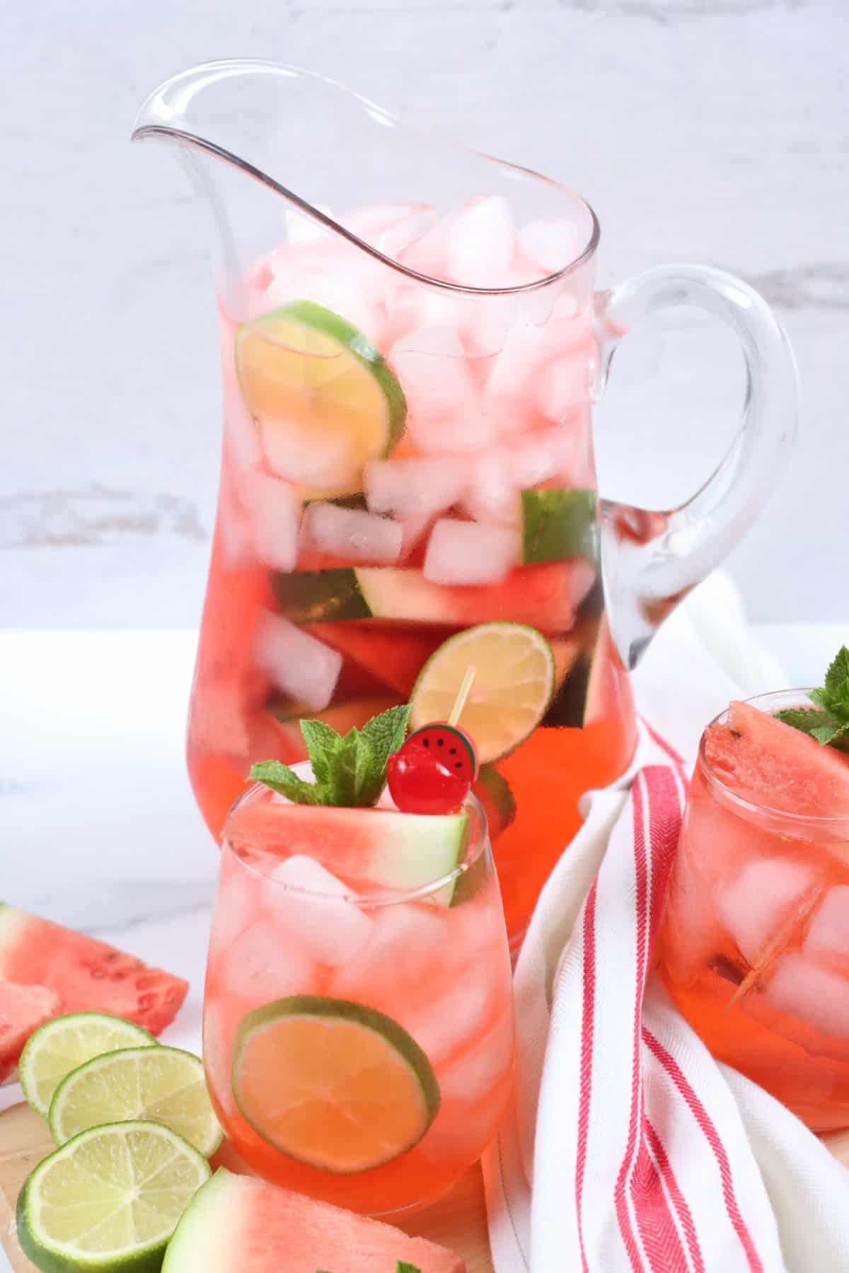 Stemless wine glass of watermelon sangria, ice cubes, fresh mint, glass pouring pitcher in background.