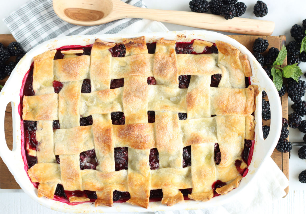 White oval baking dish with cobbler with pie crust on top, fresh blackberries around.