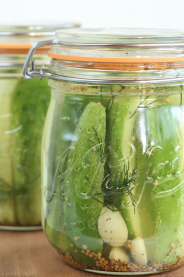 Dill pickle spears in clear glass jar with clasp lid and rubber orange seal on small wooden cutting board.