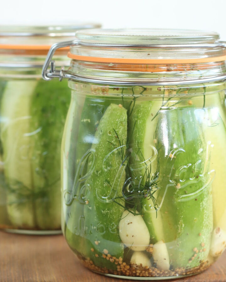Dill pickle spears in clear glass jar with clasp lid and rubber orange seal on small wooden cutting board.