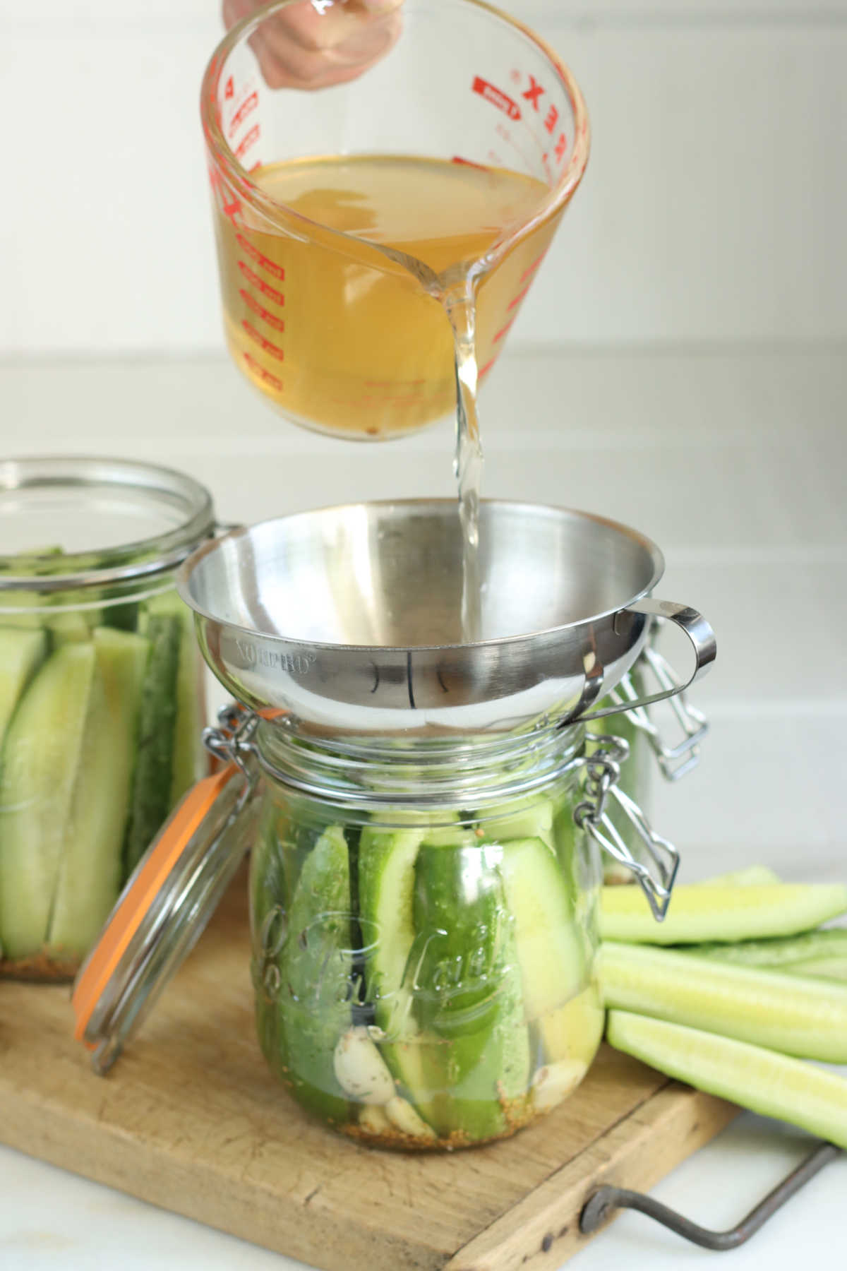 Pouring pickling brine over dill pickles in glass jar on wooden cutting board.