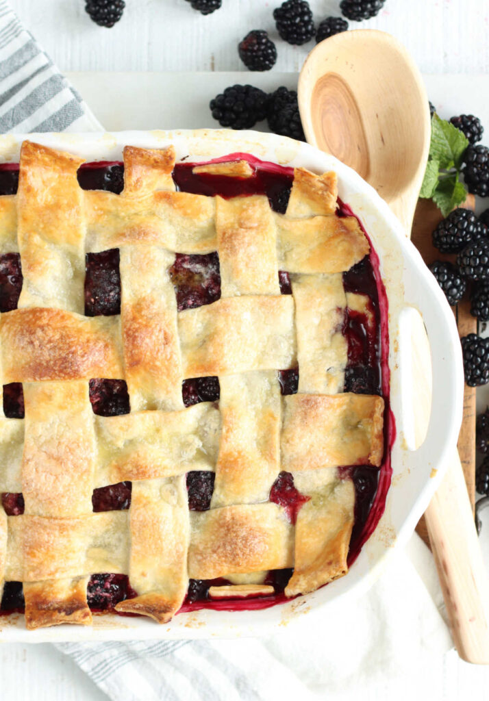 Blackberry cobbler with lattice pie crust in white baking dish, wooden spoon on right.