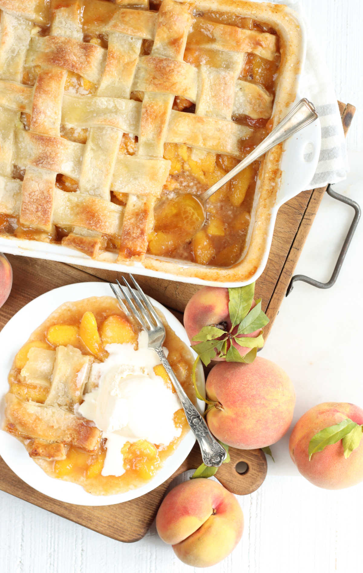 Peach cobbler with pie crust in white baking dish, piece on small white plate and fork, peaches around.