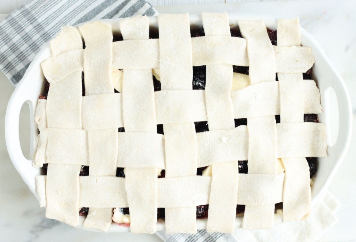 Lattice pie crust on top of cobbler in white rectangle baking dish on white marble.