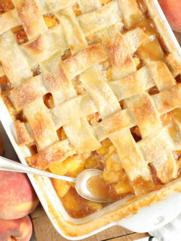 Peach cobbler with lattice pie crust, spoon in baking dish, fresh peaches around.
