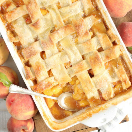 Peach cobbler with lattice pie crust, spoon in baking dish, fresh peaches around.